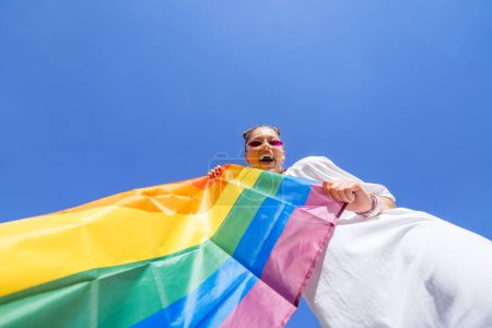 Téléchargez les photos : Une jeune femme développe un drapeau arc-en-ciel contre le ciel pour soutenir la communauté LGBT - en image libre de droit