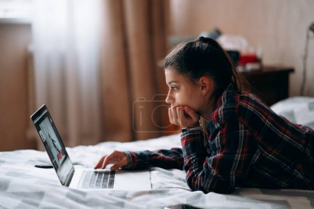 Téléchargez les photos : Concentration sélective de la jeune femme travaille avec un ordinateur portable couché sur le lit - en image libre de droit