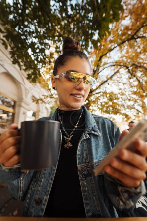 Téléchargez les photos : Jeune femme assise à l'extérieur café sur la table à boire boisson tenant dans la main smartphone, en utilisant la recherche de téléphone portable - en image libre de droit