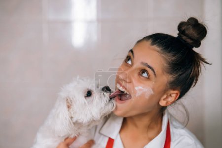 Foto de Sonriente joven en la cocina sosteniendo un lindo perro maltés blanco - Imagen libre de derechos