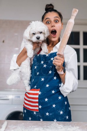 Photo for Cheerful woman with a rolling pin holding a cute white Maltese dog in the kitchen - Royalty Free Image