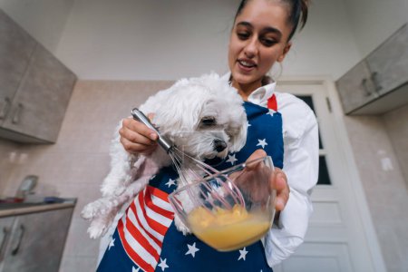 Photo for A smiling young woman is holding a cute white maltese dog while whisking an egg in the kitchen - Royalty Free Image