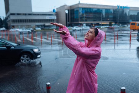 Foto de Young smiling woman with a pink raincoat on the street while enjoying a walk through the city on a rainy day. - Imagen libre de derechos