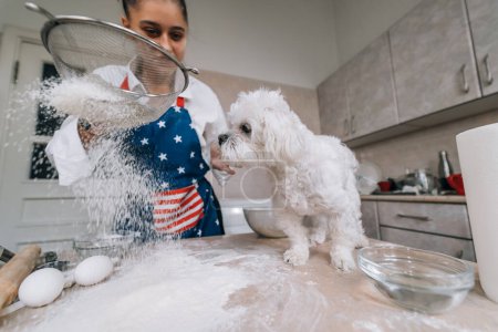 Photo for Young woman in the kitchen sifts flour together with a dog - Royalty Free Image
