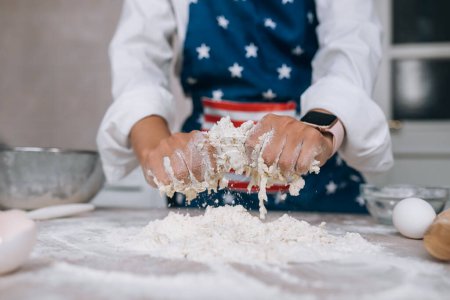 Téléchargez les photos : Le processus de fabrication de la pâte pour un repas close-up. Une jeune femme au foyer dans un tablier pétrit la pâte avec ses mains. - en image libre de droit