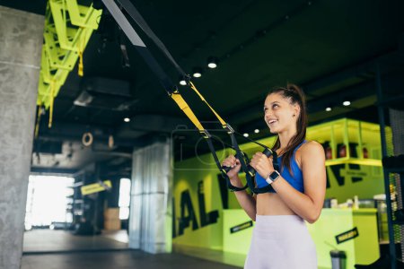 Foto de Mujer atlética joven con cuerpo perfecto en ropa deportiva haciendo ejercicios con correas de fitness trx en el gimnasio - Imagen libre de derechos