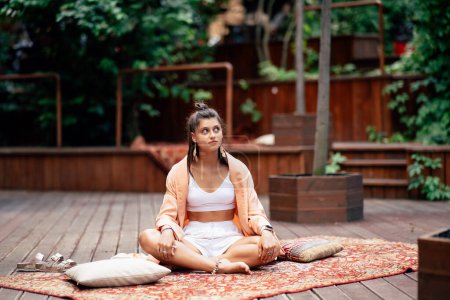 Téléchargez les photos : Jeune femme pratique le yoga assis sur un podium en bois dans le jardin le matin ensoleillé - en image libre de droit
