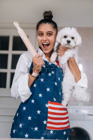 Téléchargez les photos : Femme gaie avec un rouleau à pâtisserie tenant un chien maltais blanc mignon dans la cuisine - en image libre de droit