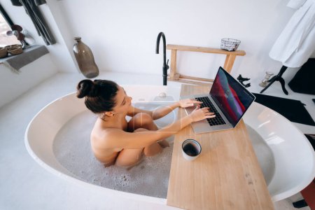 Foto de Mujer joven que trabaja en el ordenador portátil mientras toma una bañera en casa - Imagen libre de derechos