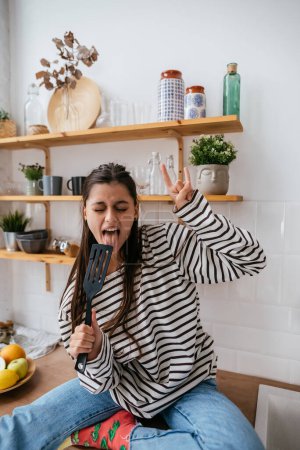 Photo for Funny beautiful woman singing into spatula, holding spatula as microphone, playful girl having fun at the kitchen. - Royalty Free Image