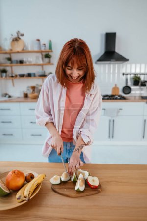 Photo for Happy healthy woman cutting fruits on a wooden board while making breakfast in a kitchen - Royalty Free Image