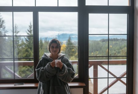 Foto de Mujer joven de pie junto a la ventana sostiene una taza y mira a la cámara - Imagen libre de derechos
