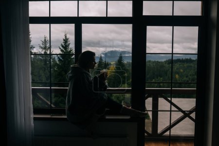 Foto de Silueta de una mujer sentada en el alféizar de la ventana con una taza y mirando las montañas y la naturaleza verde - Imagen libre de derechos