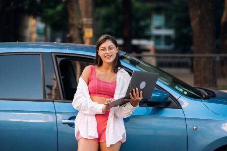 Téléchargez les photos : A young woman uses a laptop near a car in city center outdoors - en image libre de droit