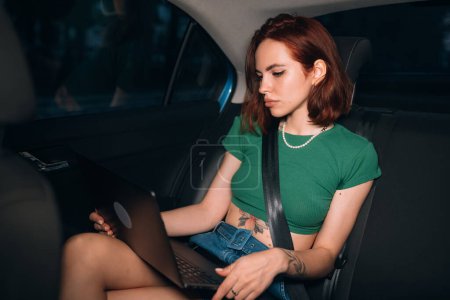 Photo for Beautiful woman is using a laptop and smiling while sitting on back seat in the car - Royalty Free Image