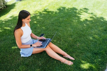 Photo for The concept of remote work. A young girl works with a laptop in the fresh air in the park, sitting on the lawn. - Royalty Free Image