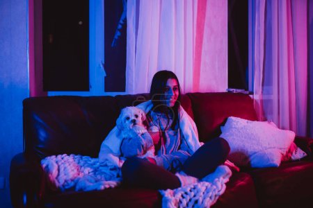 Téléchargez les photos : Belle jeune fille à la maison dans une pièce sombre jouant avec son chien et s'assoit devant un moniteur ou une télévision. Photo de haute qualité - en image libre de droit