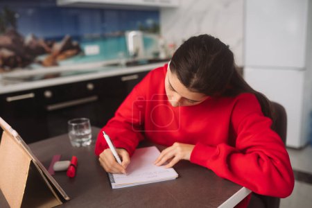 Foto de Hermosa chica con auriculares está sosteniendo un bolígrafo en su mano y escribir información de la tableta a un cuaderno. Foto de alta calidad - Imagen libre de derechos