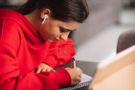 Foto de Mujer con auriculares está sosteniendo un bolígrafo en la mano y escribiendo información de la tableta a un cuaderno. Foto de alta calidad - Imagen libre de derechos
