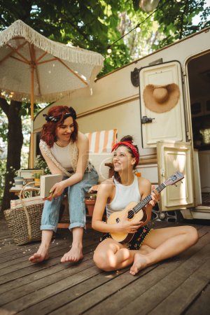 Foto de Chicas hippies felices están teniendo un buen rato con tocar en la guitarra en caravana remolque. Vacaciones, vacaciones, concepto de viaje.Foto de alta calidad - Imagen libre de derechos