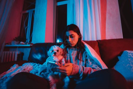 Photo for Beautiful young girl at home in a dark room with smartphone in her hands playing with her dog and sits in front of a monitor or TV. High quality photo - Royalty Free Image