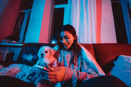 Foto de Hermosa joven en casa en una habitación oscura con teléfono inteligente en las manos jugando con su perro y se sienta frente a un monitor o TV. Foto de alta calidad - Imagen libre de derechos
