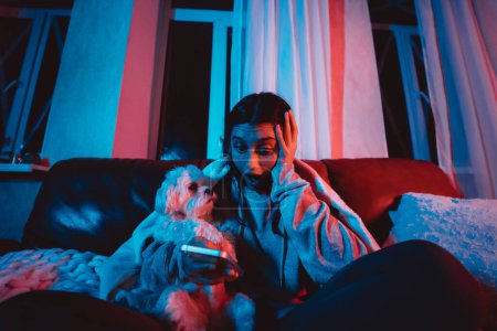 Foto de Hermosa joven en casa en una habitación oscura jugando con su perro y se sienta frente a un monitor o TV. Foto de alta calidad - Imagen libre de derechos