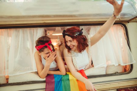 Foto de Retrato de una linda pareja de lesbianas. Dos chicas pasan tiempo tiernamente juntas viendo películas en un portátil en un remolque de caravana con bandera LGBT en la pared. Amor y actitud. Concepto LGBT. Foto de alta calidad - Imagen libre de derechos