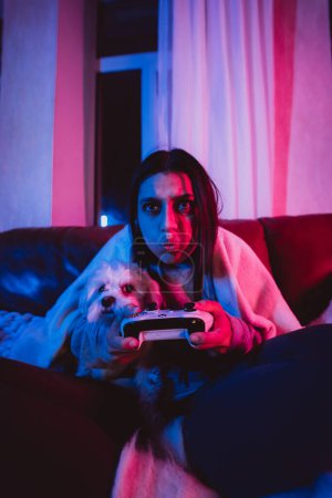 Téléchargez les photos : Gros plan portrait d'une gamer ou d'une streamer girl à la maison dans une pièce sombre avec une manette de jeu jouant avec son chien et assise devant un moniteur ou une télévision. Photo de haute qualité - en image libre de droit