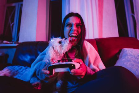 Foto de Primer plano retrato de un jugador o una chica streamer en casa en una habitación oscura con un controlador de juego jugando con su perro y se sienta delante de un monitor o TV. Emociones locas. Foto de alta calidad - Imagen libre de derechos