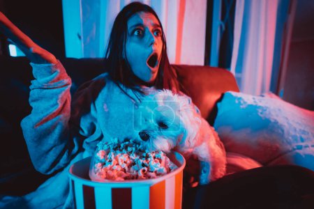 Photo for Close up portrait of Beautiful young girl at home in a dark room with a popcorn bucket playing with her dog and sits in front of a monitor or TV watching movie. Crazy emotions. High quality photo - Royalty Free Image