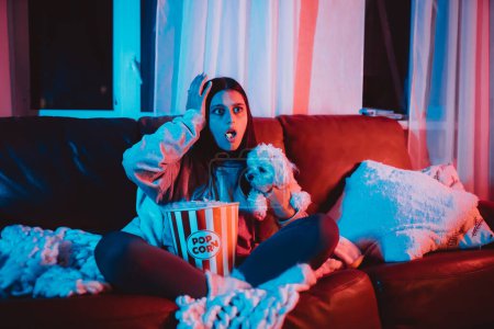 Photo for Beautiful young girl watching movie at home in a dark room with a popcorn bucket playing with her dog and sits in front of a monitor or TV watching movie. Crazy emotions. High quality photo - Royalty Free Image