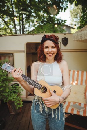 Happy hippie girl is having a good time with playing on guitar in camper trailer. Holiday, vacation, trip concept.High quality photo