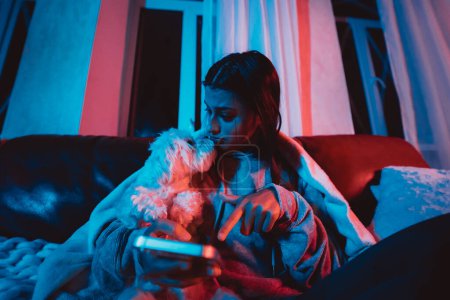 Téléchargez les photos : Belle jeune fille à la maison dans une pièce sombre jouant avec son chien et s'assoit devant un moniteur ou une télévision. Photo de haute qualité - en image libre de droit