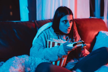 Photo for A gamer or a streamer girl at home in a dark room with a game controller and popcorn bucket playing with her dog and sits in front of a monitor or TV. High quality photo - Royalty Free Image