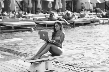 Photo for Cute young woman sunbathing sitting in front of the pool, working with a laptop at summer day. Black and white photo - Royalty Free Image