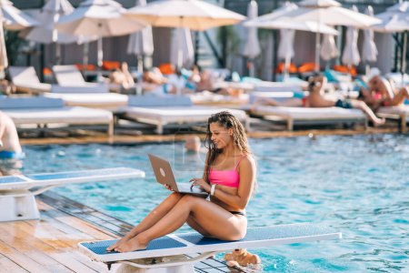 Téléchargez les photos : Jolie jeune femme bronzant assis devant la piscine, travaillant avec un ordinateur portable le jour de l'été - en image libre de droit