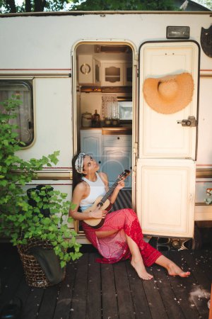 Foto de Chica hippie feliz están teniendo un buen rato con tocar en la guitarra en caravana remolque. Vacaciones, vacaciones, concepto de viaje.Foto de alta calidad - Imagen libre de derechos