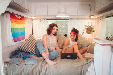 Photo for Portrait of a cute lesbian couple. Two girls spend time tenderly together watching movie on laptop in a camper trailer with LGBT flag on the wall. Love and attitude. LGBT concept. High quality photo - Royalty Free Image