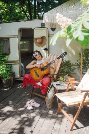 Glückliche Hippie-Mädchen haben eine gute Zeit mit dem Spielen auf der Gitarre im Wohnwagen. Urlaub, Urlaub, Reisekonzept. Hochwertiges Foto