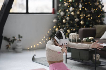 Foto de Una joven de moda en atuendo deportivo, luciendo un auricular de realidad virtual, hace yoga con un árbol de Navidad en el fondo. Foto de alta calidad - Imagen libre de derechos