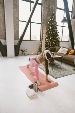 Photo for The stylish young woman, in her workout outfit and a virtual reality headset, does yoga poses beside a Christmas tree. High quality photo - Royalty Free Image