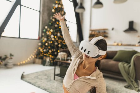 Photo for A fitness-oriented young woman practices yoga poses while wearing VR glasses by a Christmas tree. High quality photo - Royalty Free Image