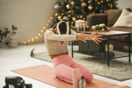 Téléchargez les photos : Une jeune femme à la mode en tenue de sport, arborant un casque de réalité virtuelle, fait du yoga avec un arbre de Noël en arrière-plan. Photo de haute qualité - en image libre de droit
