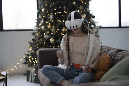 Foto de Absorto de la comodidad de un ambiente navideño en casa, una mujer joven y elegante se pone un auricular de realidad virtual. Foto de alta calidad - Imagen libre de derechos