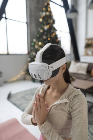 Téléchargez les photos : À l'aide d'un casque de réalité virtuelle, une jeune femme en tenue de sport rose pratique le yoga près d'un arbre de Noël. Photo de haute qualité - en image libre de droit
