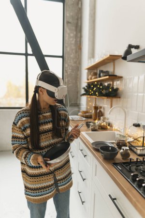 Téléchargez les photos : Une belle fille élégante cuisine selon une recette de Noël dans un casque de réalité virtuelle dans la cuisine de vacances. Photo de haute qualité - en image libre de droit