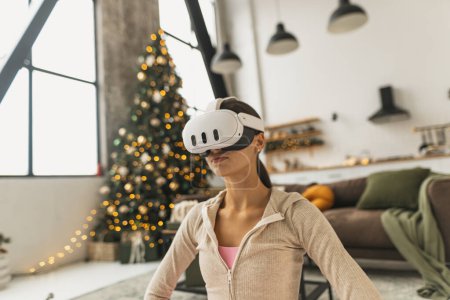 Photo for A young woman, wearing a pink sports outfit, practices yoga with a virtual reality headset near a Christmas tree. High quality photo - Royalty Free Image