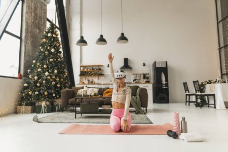 Photo for A pleasant young lady does stretching exercises beside a Christmas tree while wearing VR glasses. High quality photo - Royalty Free Image