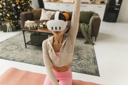 Foto de Una joven encantadora, con gafas VR, está haciendo ejercicios de estiramiento junto al árbol de Navidad. Foto de alta calidad - Imagen libre de derechos